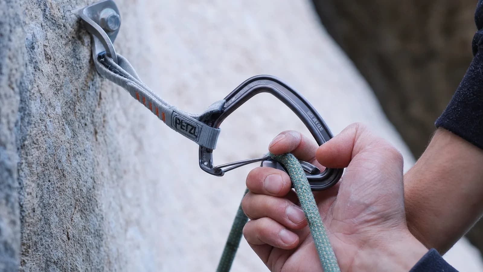 Un grimpeur en tête en plein clip de dégaine sur une voie en dévers léger, main droite poussant la corde dans un mousqueton orange Petzl Ange, bras gauche tendu sur un bac jaune