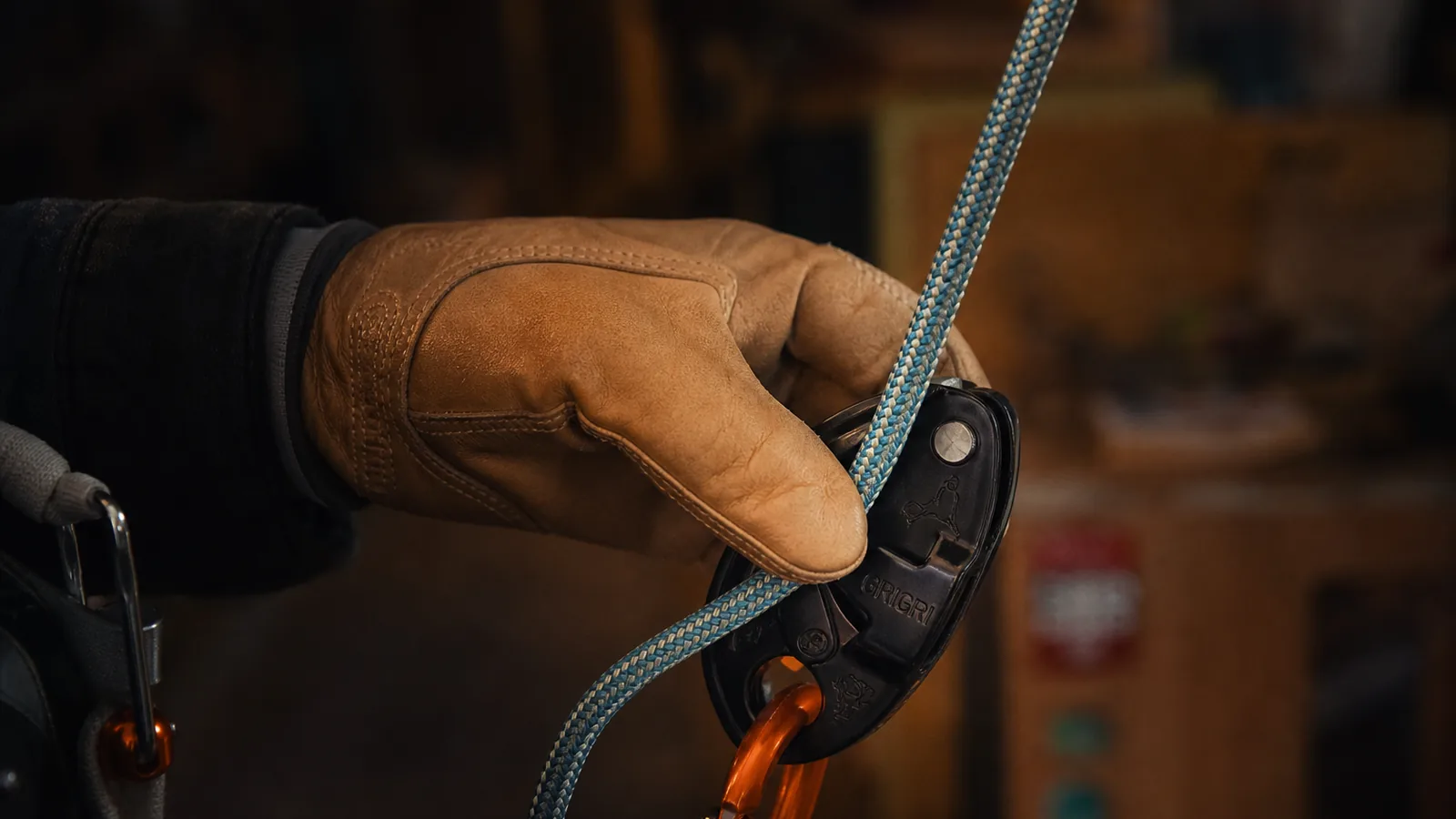 Paire de gants d'assurage en cuir nubuck accrochés à un baudrier par un mousqueton, vue de près en lumière naturelle, sangle de poignet visible, paume légèrement marquée par l'usage