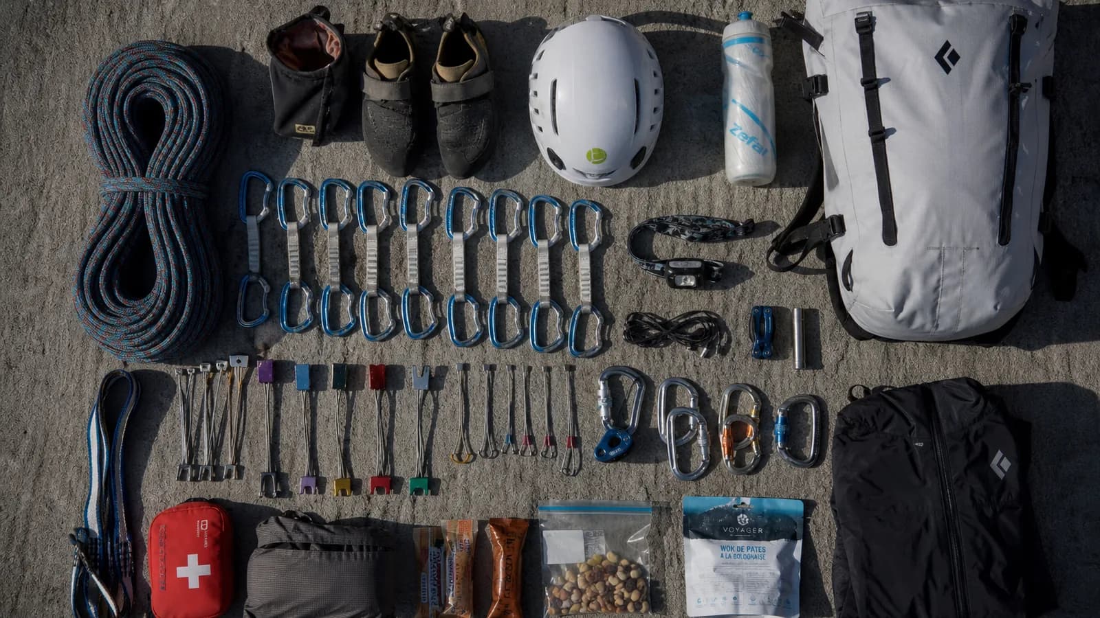 Sac d'escalade ouvert au sol au pied d'une falaise calcaire, matériel étalé sur un rocher : corde 70 m enroulée, dégaines colorées alignées, casque rouge, sangles Dyneema, lumière matinale dorée