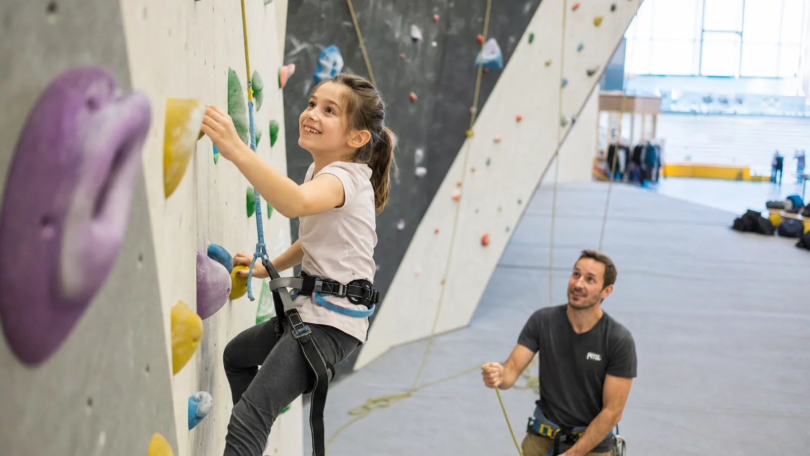Petite fille en baudrier rouge et chaussons gris en train de grimper une voie de salle facile, coach souriant qui assure depuis le sol, prises colorées à sa portée